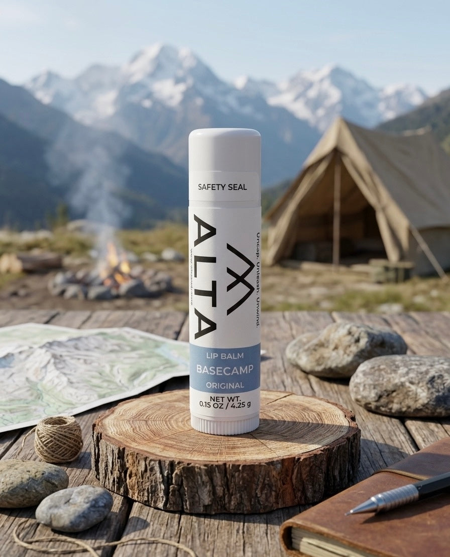 A tube of ALTA Basecamp Original Lip Balm standing on a wooden stump with a mountain landscape, a canvas tent, and a campfire in the background.