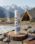 A tube of ALTA Basecamp Original Lip Balm standing on a wooden stump with a mountain landscape, a canvas tent, and a campfire in the background.