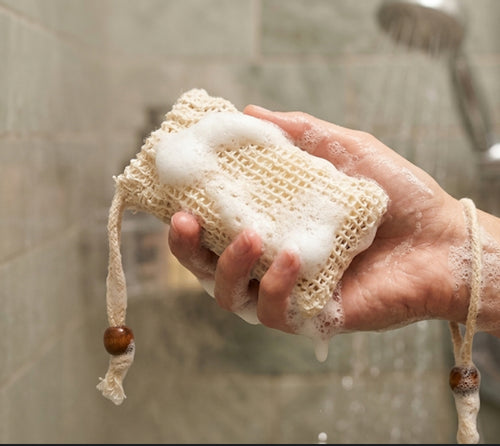 Hand holding a sudsy ALTA Bar Sleeve made of natural sisal fibers for exfoliating skin and extending bar soap life in a shower setting.