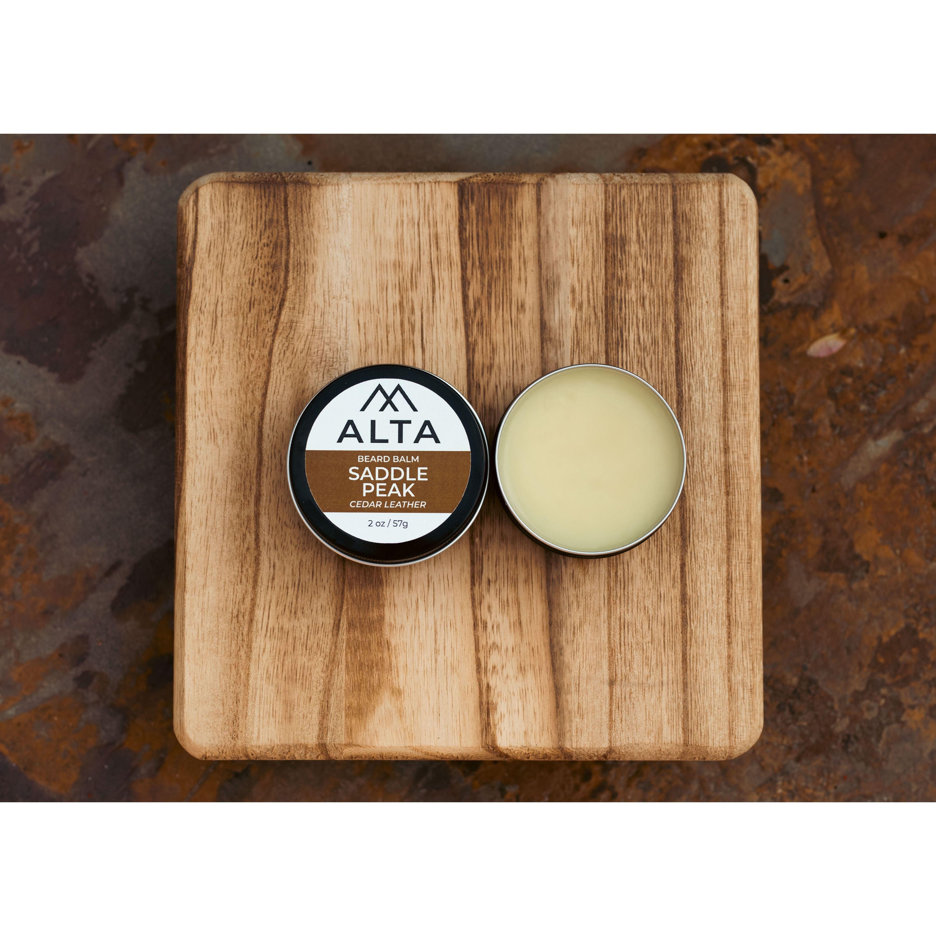 Alta Body Saddle Peak Premium Beard Balm in a 2oz black tin resting on a wooden surface. Natural cedar and leather scent.